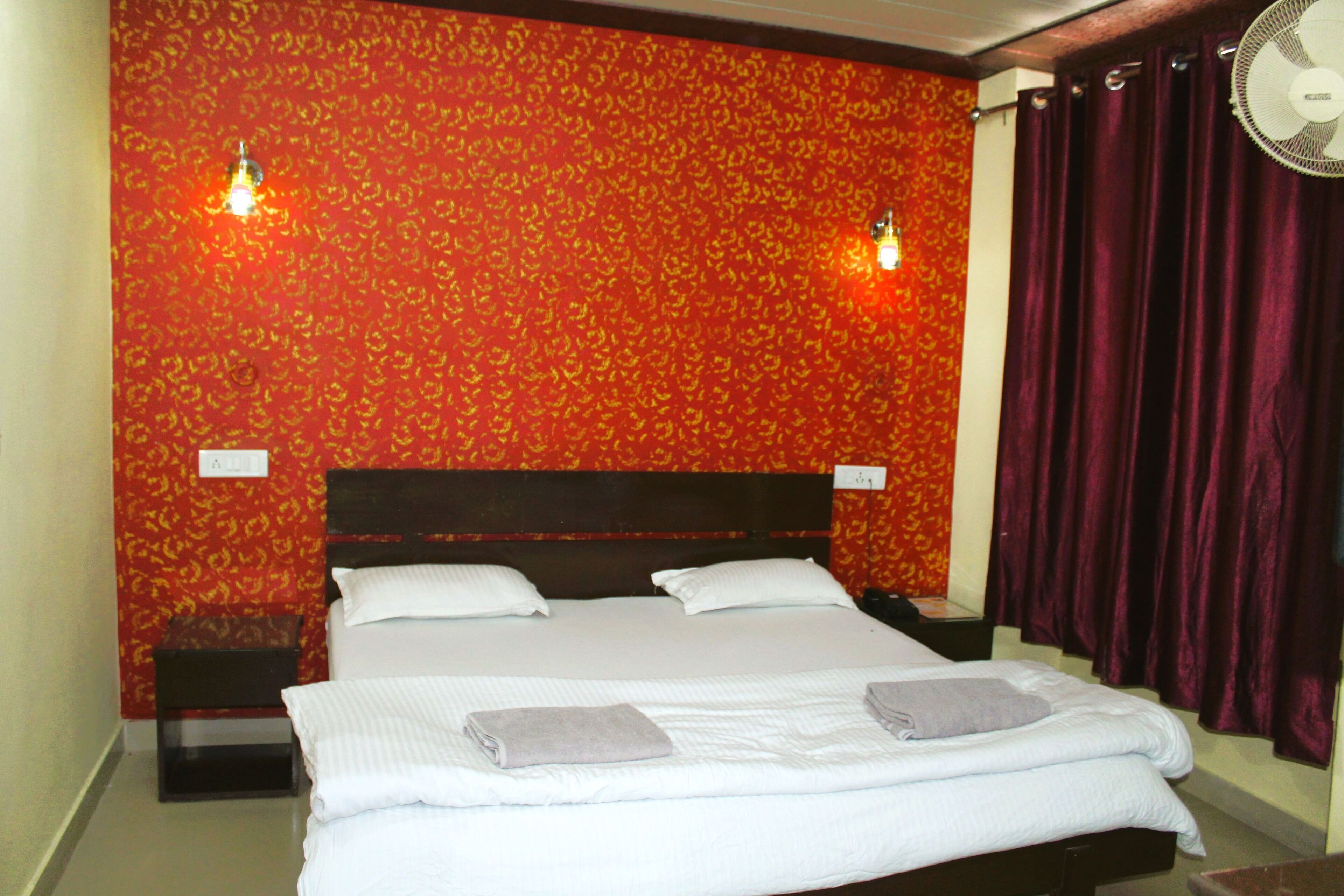 A hotel room with a bed, two bedside tables, a wall-mounted fan, and a red patterned wall.