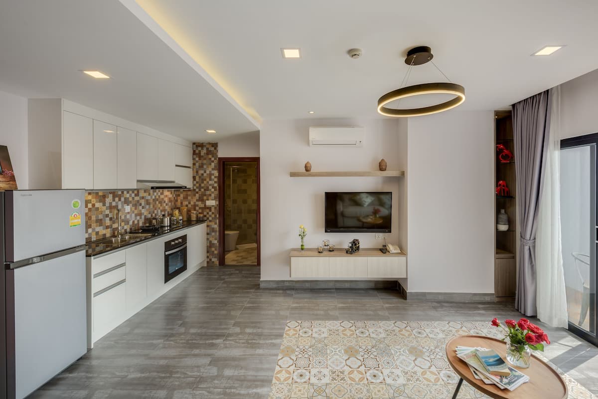 A modern kitchen with white cabinets, a built-in oven, and a refrigerator. There is a dining area with a round table and chairs, a TV mounted on the wall, and a patterned rug on the floor.