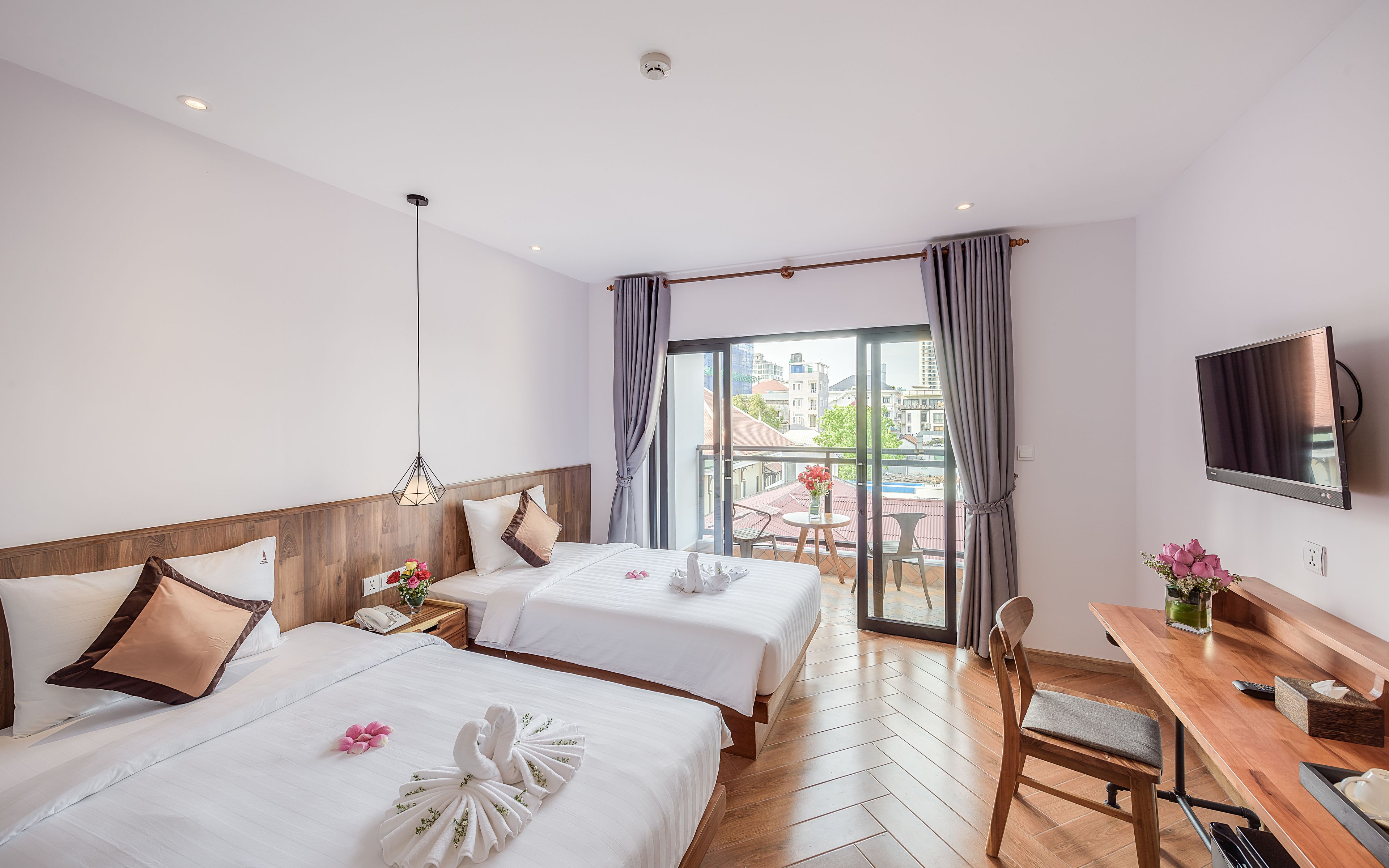 A hotel room with two beds, a dining table, a TV, and a balcony with a view of buildings.