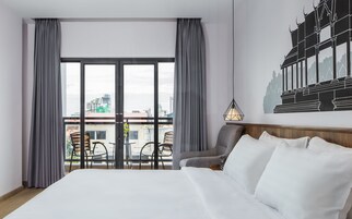 Habitación de hotel con una cama grande, cortinas grises, vista a la ciudad, una lámpara de pared y una silla junto a la ventana.