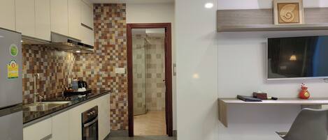 A modern kitchen with a tiled backsplash, a built-in oven, and a sink. There's a wooden door leading to a bathroom. A flat-screen TV is mounted on the wall above a desk with a chair.