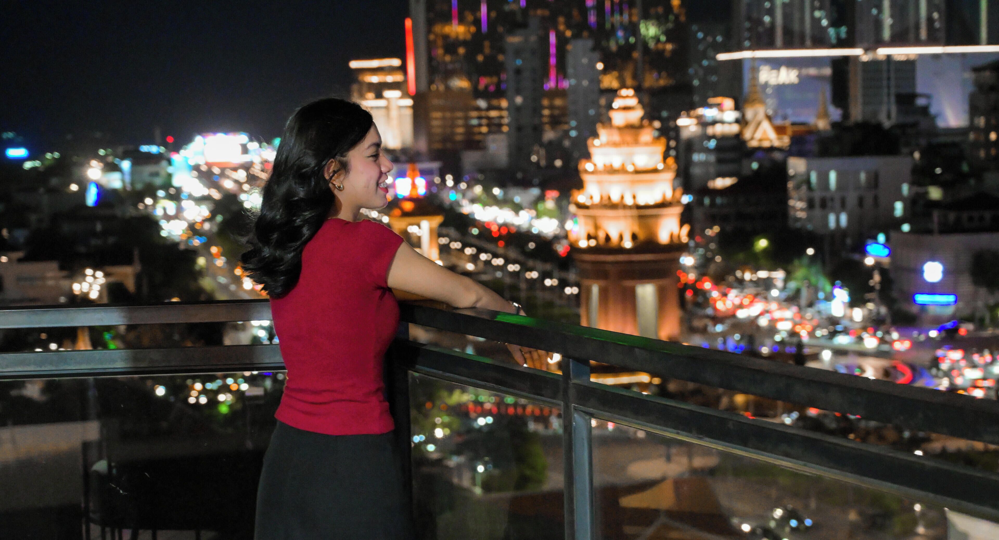 Une femme se tient près d’une balustrade, d’où elle peut admirer un paysage urbain avec des bâtiments illuminés et une structure ressemblant à une pagode.
