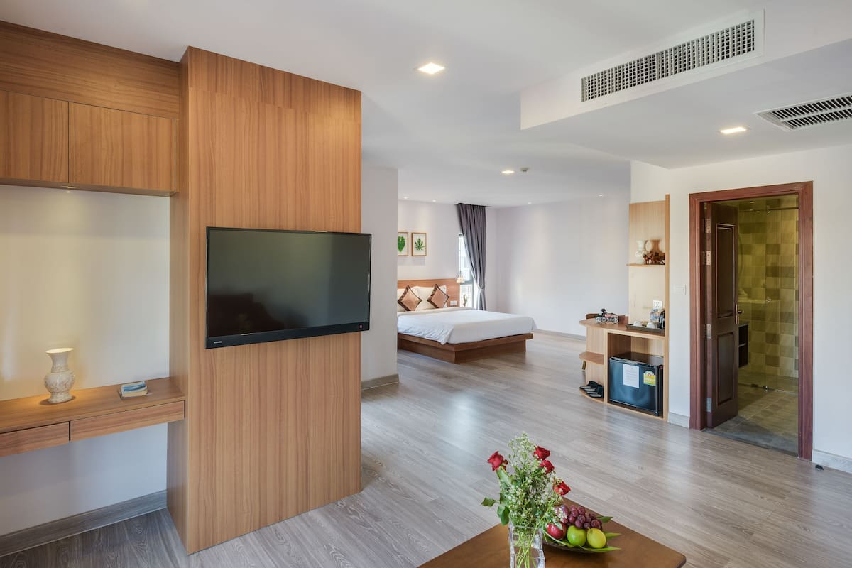 A modern hotel room with a wooden cabinet, a flat-screen TV, a bed with a headboard, a desk with a chair, a small table with a vase of flowers, and a shower area.