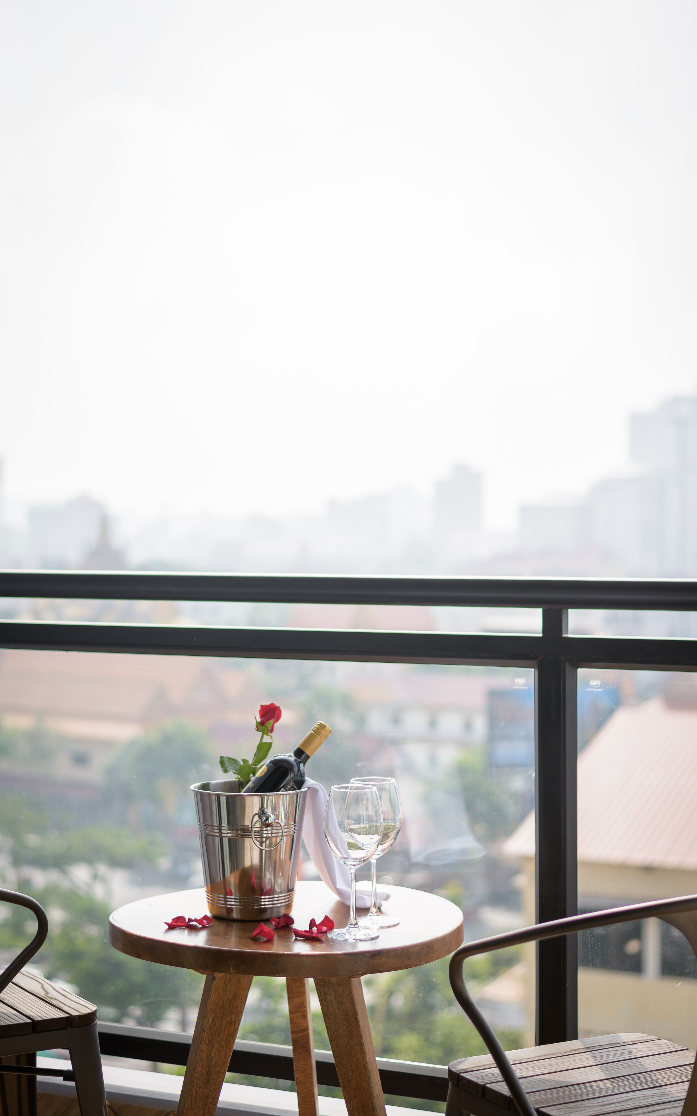 A table with a champagne bottle, glasses, and a rose on a balcony overlooking a cityscape.