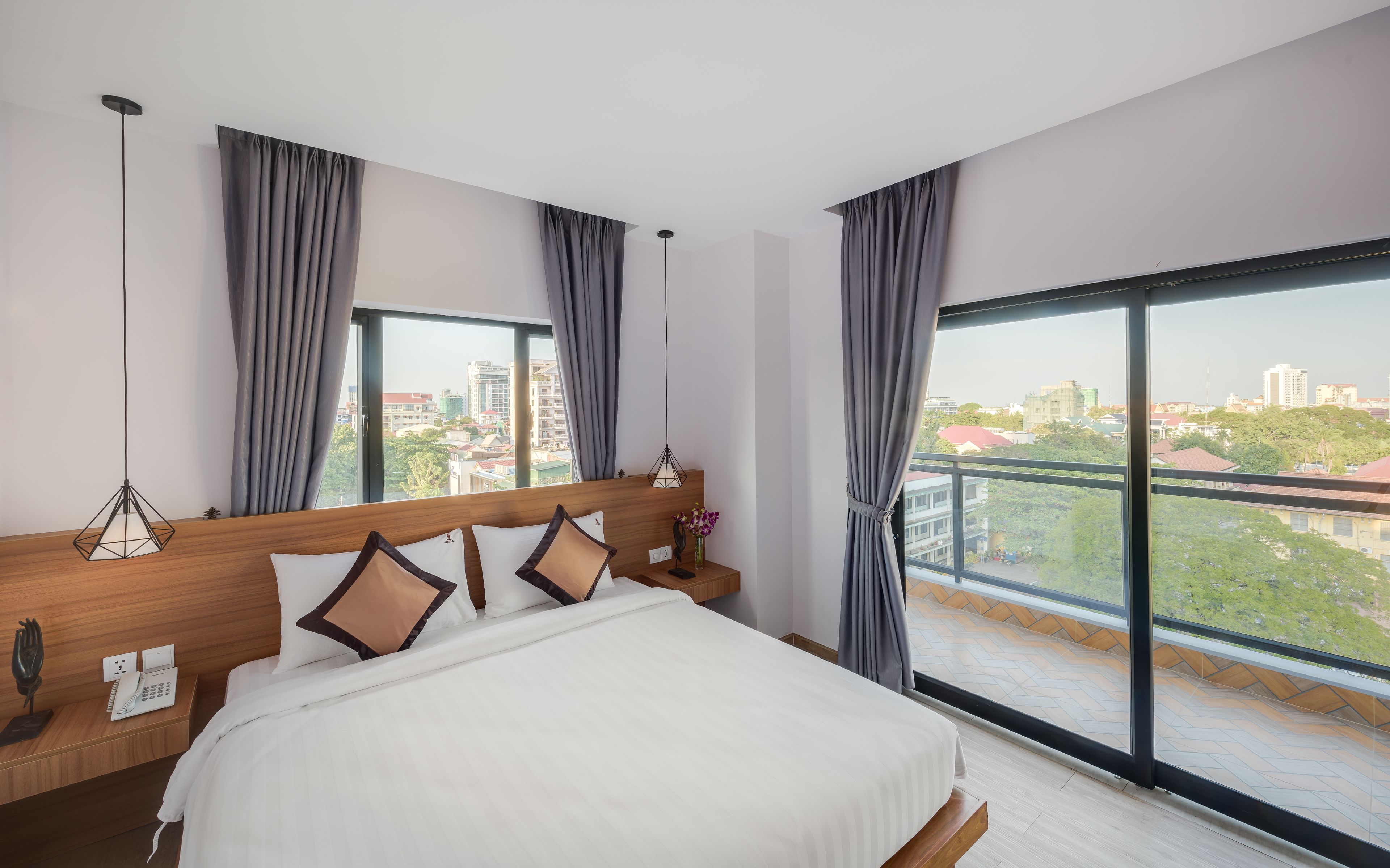 Una habitación de hotel moderna con una cama grande, una mesita de noche con un teléfono y una lámpara, vistas a la ciudad a través de amplias ventanas y cortinas grises.