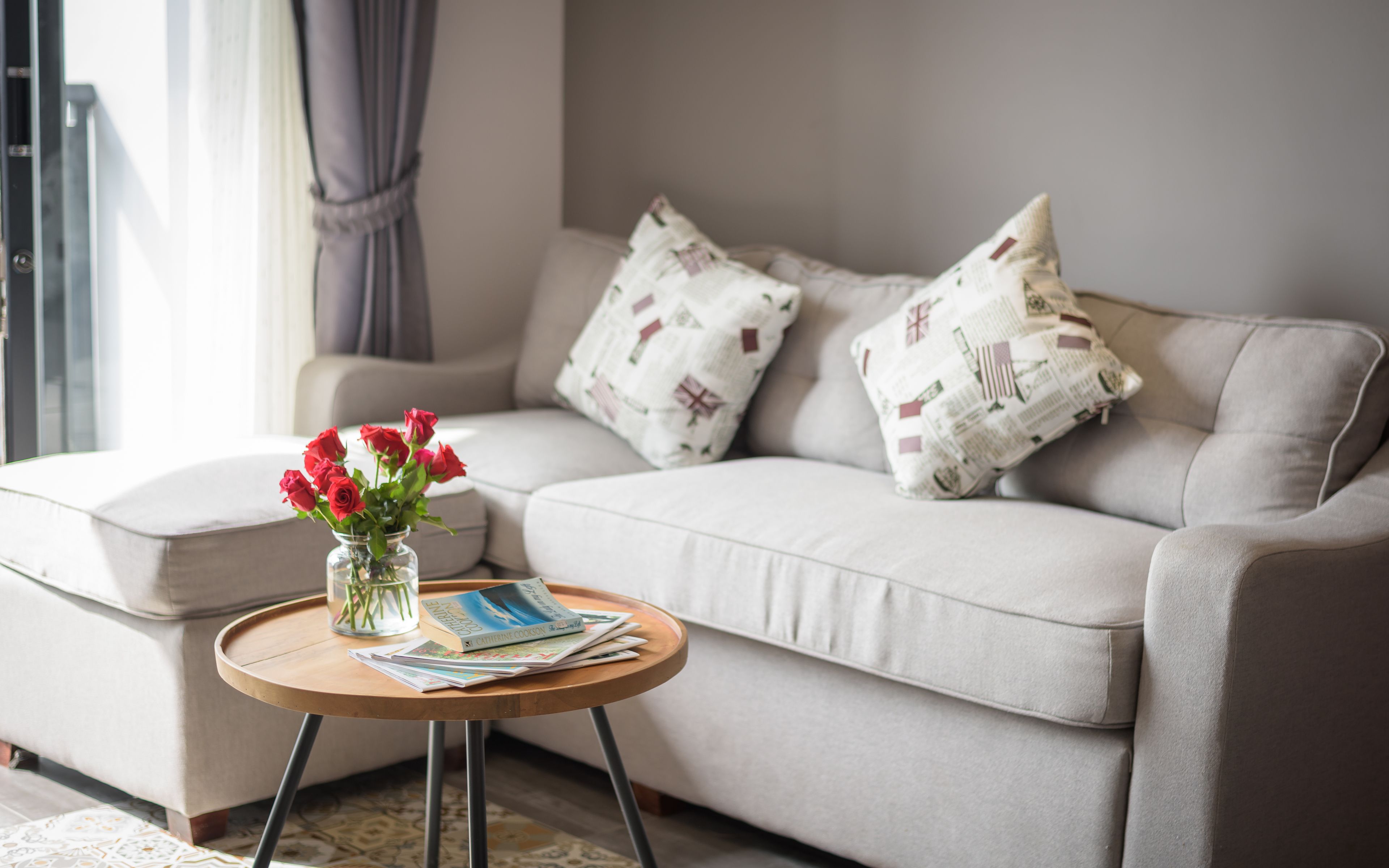 Ein Wohnzimmer mit einem beigen Sofa, einem runden Holztisch, einer Vase mit roten Blumen und gemusterten Kissen.