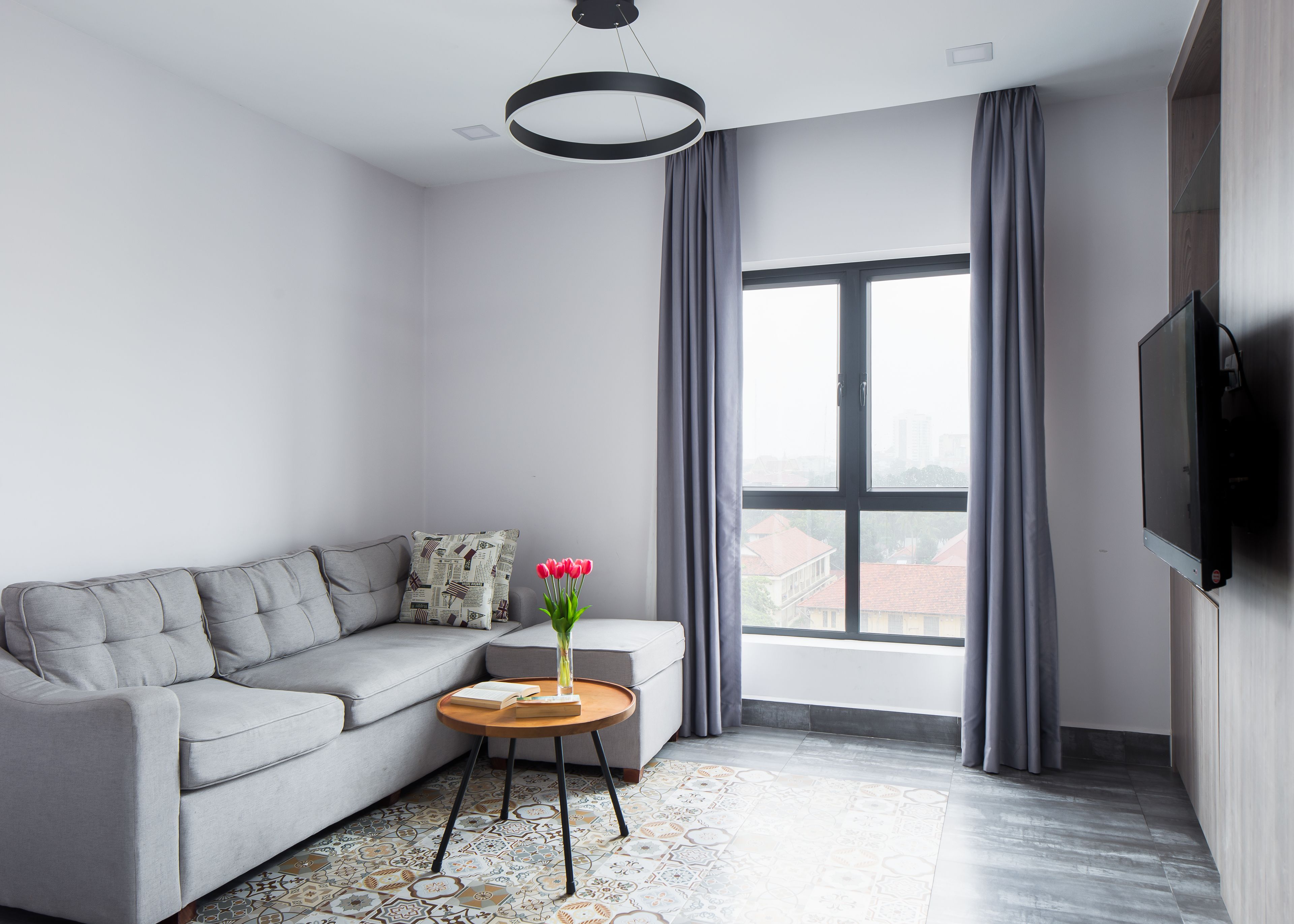 Una sala de estar moderna con un sofá gris, una mesita redonda con un libro y un jarrón de flores, un ventanal con cortinas y un televisor de pantalla plana montado en la pared.
