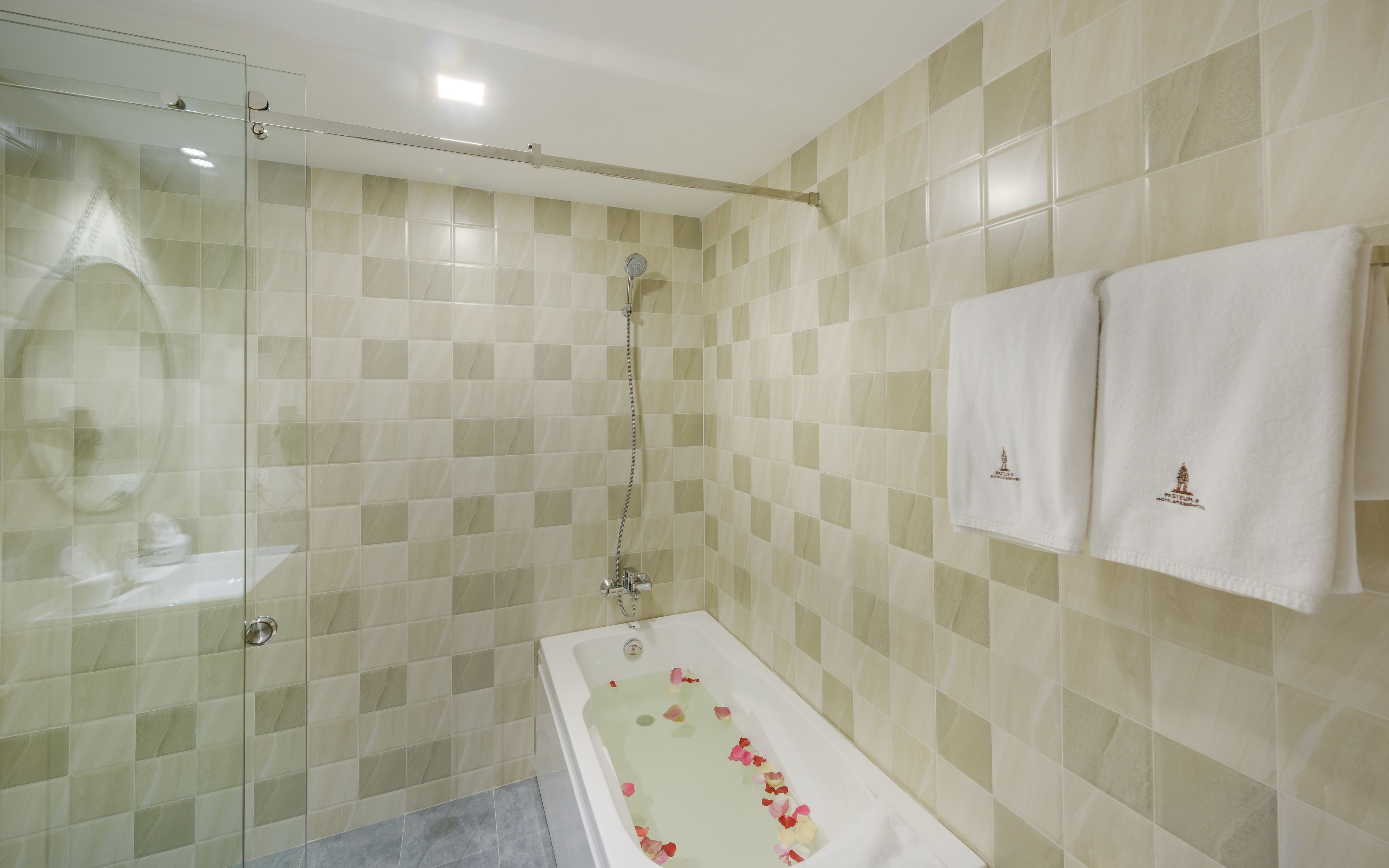 Une salle de bain avec une douche à parois de verre, une baignoire remplie de pétales de rose et deux serviettes ornées d’un logo.