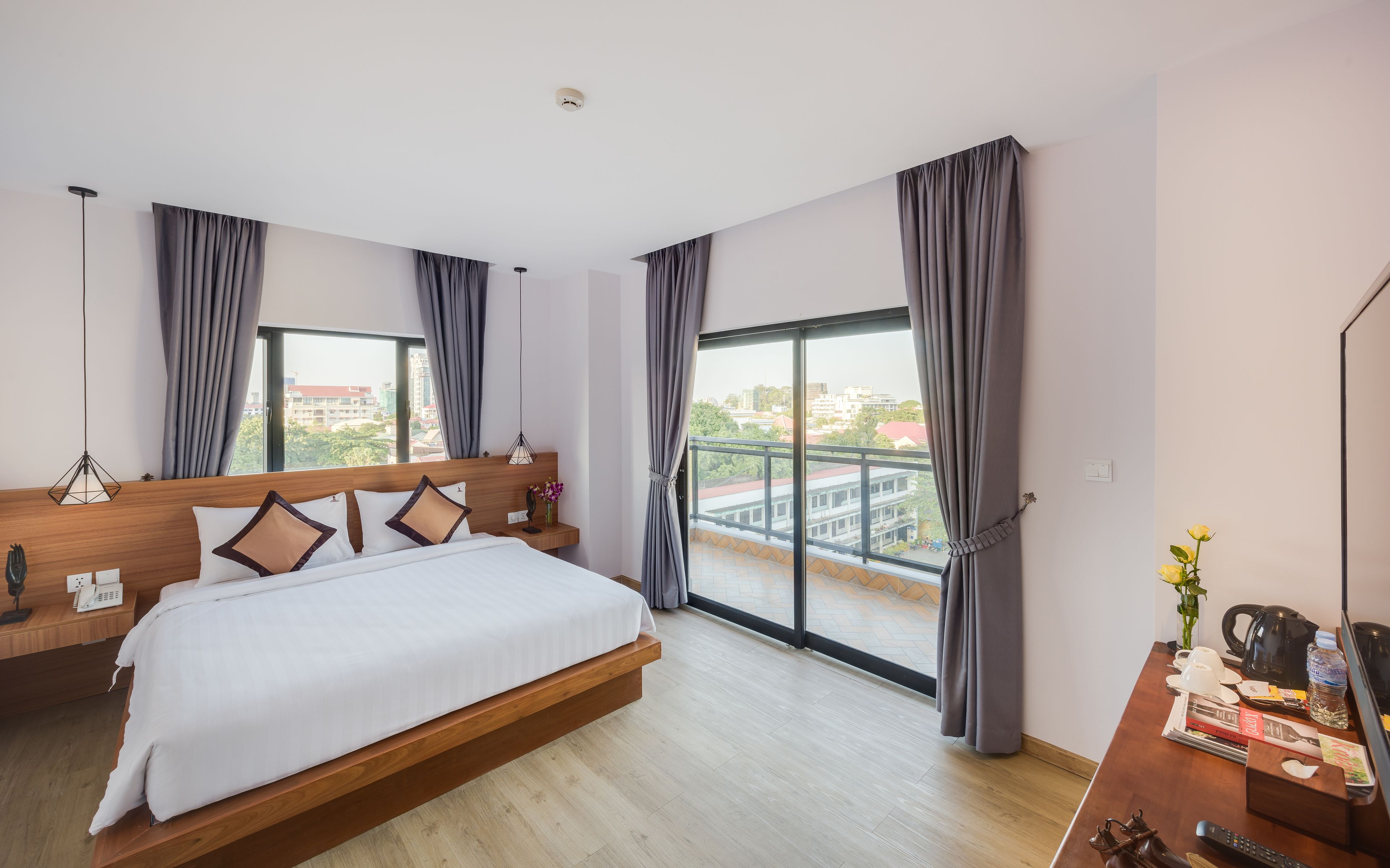 Una habitación de hotel moderna con una cama grande, un cabecero de madera, un escritorio con cafetera y vistas a la ciudad a través de amplios ventanales.