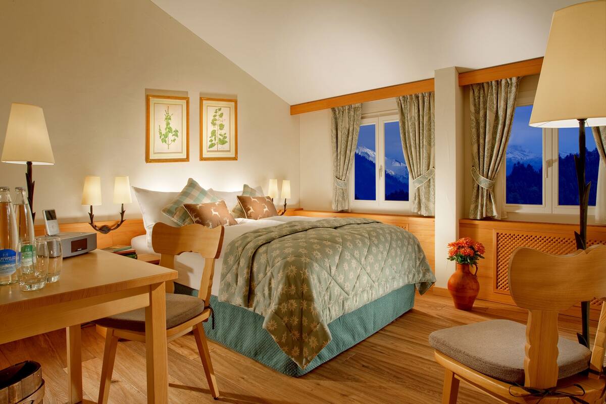 Une chambre d’hôtel comprenant un lit, des lampes de chevet, un bureau avec une chaise, une table, un vase avec des fleurs et une fenêtre avec des rideaux.