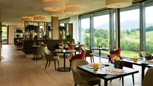 A spacious dining area with multiple tables and chairs, large windows offering a view of a green landscape, and a modern interior design with wooden flooring and pendant lighting. - Bürgenstock Hotels & Resort – Taverne 1879 (Ennetbuergen)