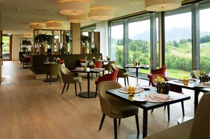 Un comedor amplio con varias mesas y sillas, ventanas grandes que ofrecen vistas a un paisaje verde, y un diseño interior moderno con piso de madera e iluminación colgante.