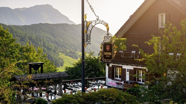 A traditional European tavern with outdoor seating, surrounded by lush greenery and mountains in the background. - Bürgenstock Hotels & Resort – Taverne 1879 (Ennetbuergen)