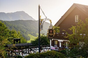 Una taberna europea tradicional con terraza al aire libre, rodeada de abundante vegetación y montañas al fondo.