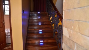 Una escalera de madera con luces LED azules en cada peldaño, que conduce a una habitación con una cama estampada y una ventana con celosía decorativa.