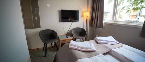 Una habitación de hotel con cama, dos sillas, escritorio, televisor y ventana con cortinas.