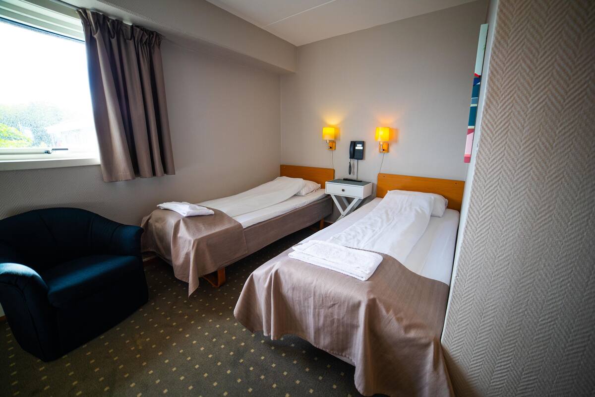 Une chambre d’hôtel avec deux lits simples, un canapé, une table de chevet et une fenêtre avec des rideaux.