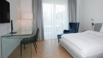 A hotel room with a bed, a desk with a chair, a chair, a lamp, and a TV on the wall.