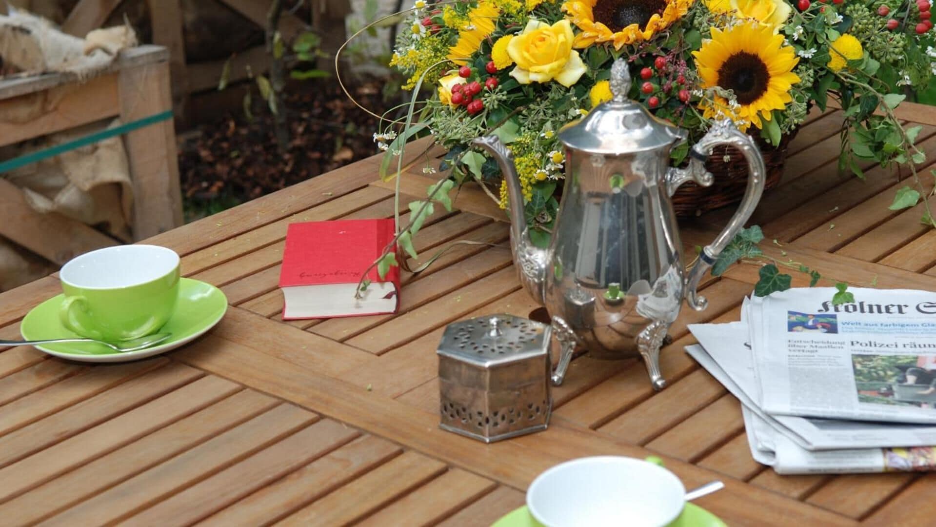 A wooden table with a green cup, a silver teapot, a grater, a red book, and a newspaper.