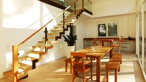 Een moderne keuken met een lange houten tafel en stoelen, een trap met glazen balustrades en een groot raam dat voor natuurlijk licht zorgt.