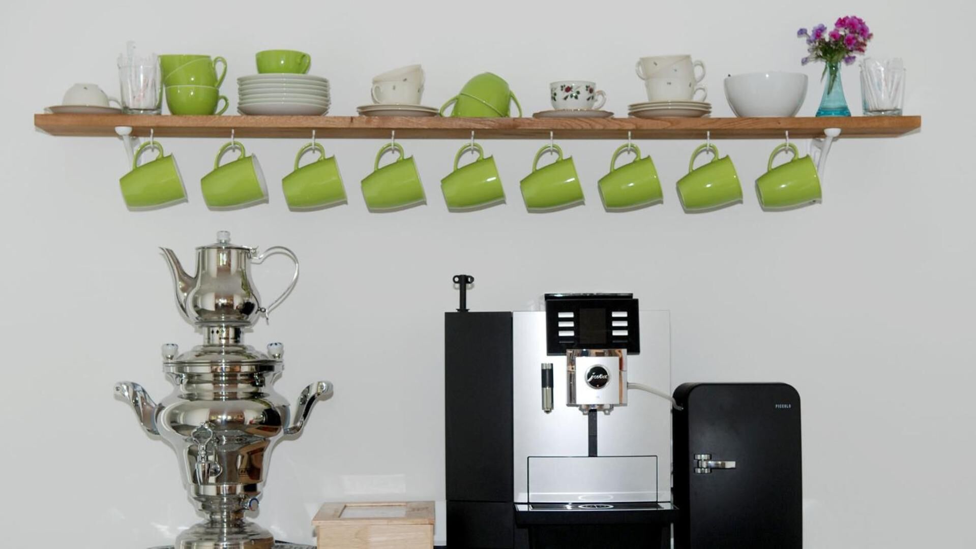 Una cocina con cafetera, tetera y una estantería con tazas y platos verdes.