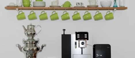 Una cocina con cafetera, tetera y una estantería con tazas y platos verdes.