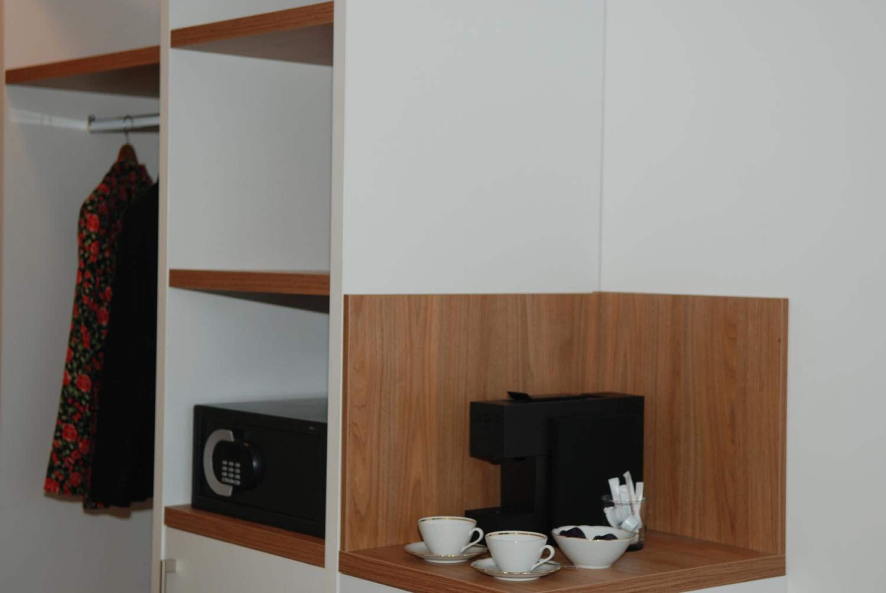 A hotel room with a wooden shelf holding cups and a coffee maker, a phone, and a black box.