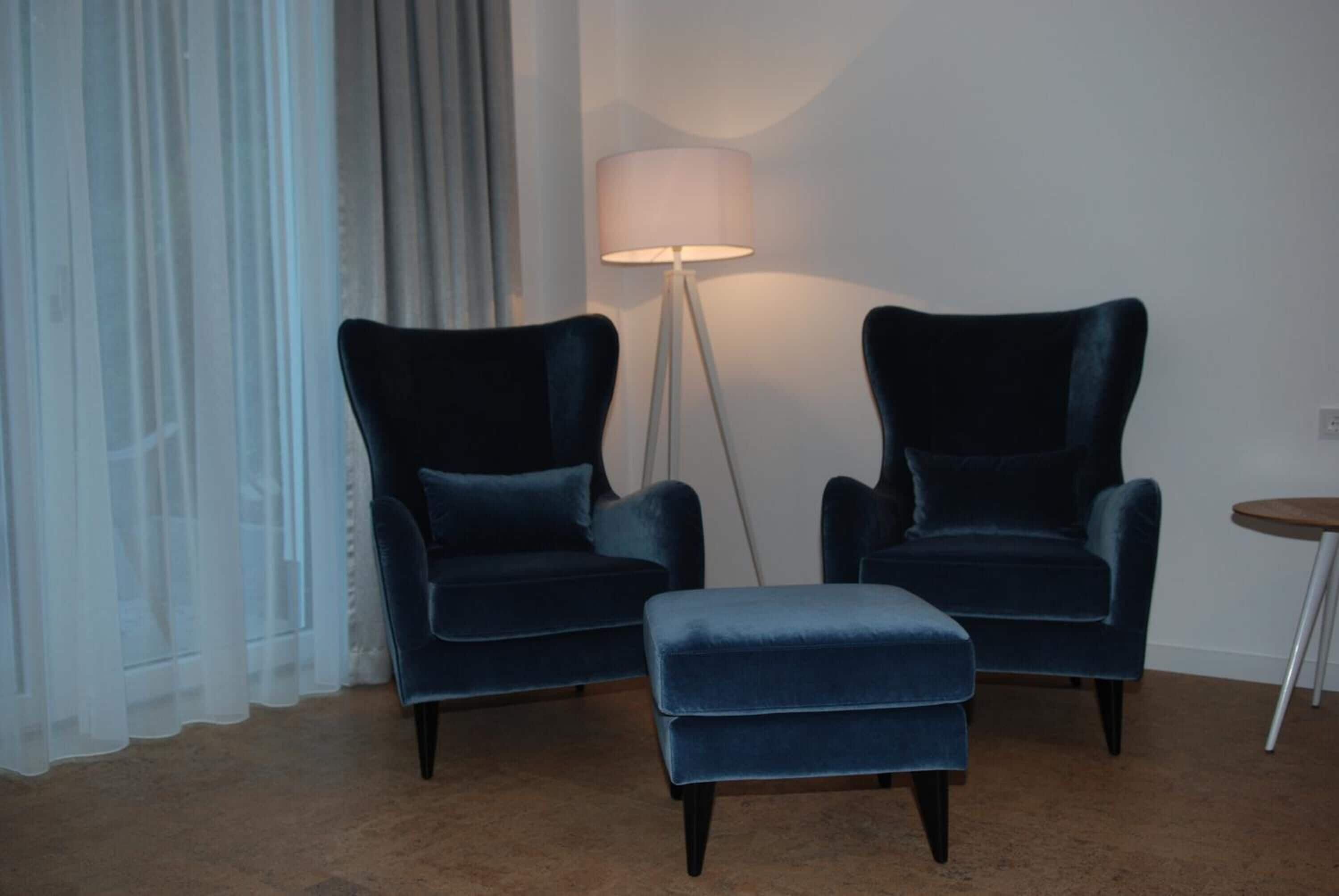 Ein Zimmer mit zwei dunkelblauen Sesseln und einem passenden Hocker, einer Stehlampe und einem kleinen Tisch.