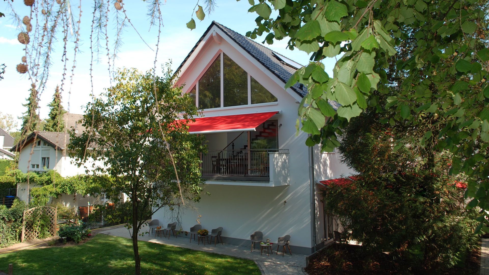Ein modernes Haus mit rotem Markisenvorbau, einem Balkon und einem Garten mit Sitzgelegenheiten.