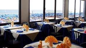 Ein Restaurant mit gedeckten Tischen, großen Fenstern mit Blick auf einen Strand und Meerblick.