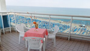 Ein Balkon mit Tisch und Stühlen, der Blick auf einen Strand mit vielen Sonnenschirmen und Menschen freigibt.