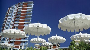Ein Hochhaus-Hotel mit mehreren Balkonen, großen weißen Sonnenschirmen und üppigem Grün im Vordergrund.