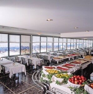 A spacious dining area with multiple tables and chairs, large windows offering a view of the city, and a buffet with various food items. - Hotel Caravelle & Minicaravelle (Jesolo)