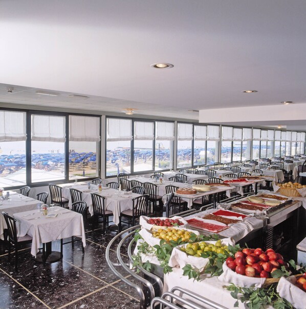 A spacious dining area with multiple tables and chairs, large windows offering a view of the city, and a buffet with various food items.