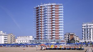 Ein hohes Hotel mit mehreren Balkonen, ein Strand mit Sonnenschirmen und ein Holzsteg entlang des Wassers.