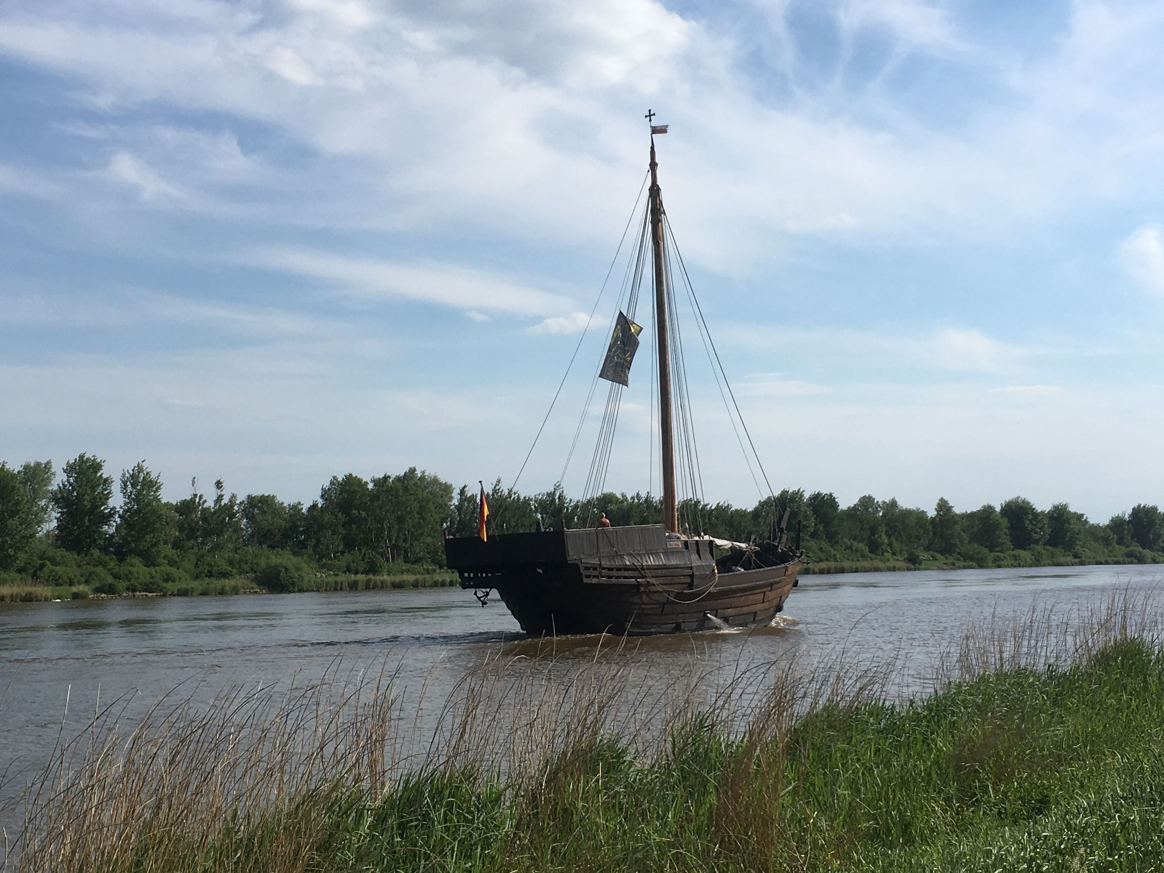 A boat on a river with a flag on top, surrounded by greenery and trees.