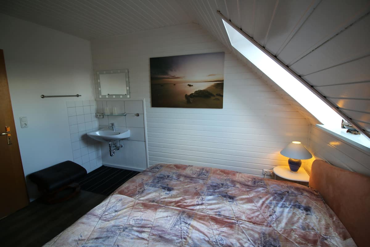 Une chambre à coucher avec un lit, une table de chevet avec une lampe, une fenêtre, une salle de bain avec un lavabo et un miroir, et un tableau accroché au mur.
