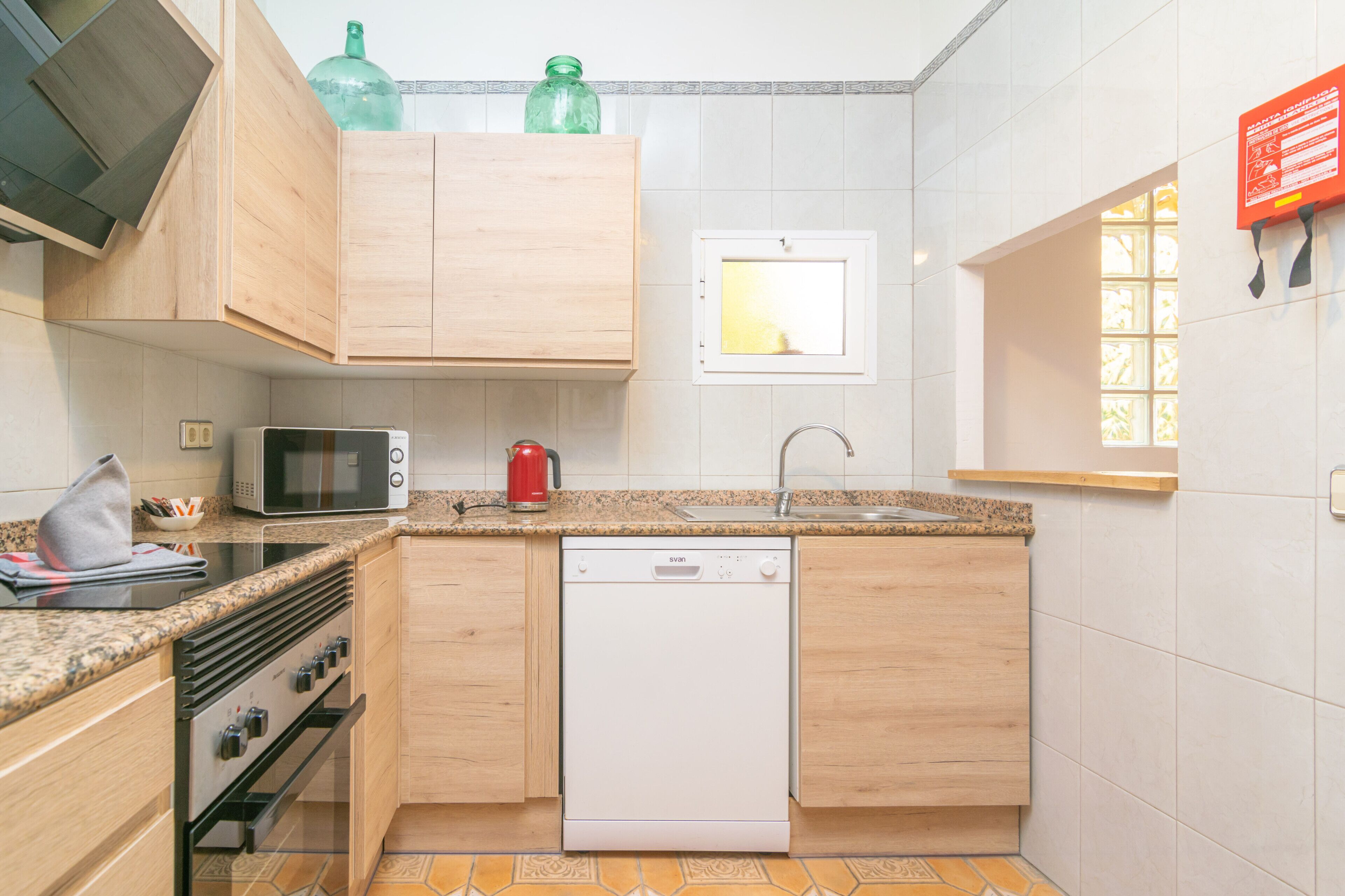 Cocina compacta con armarios de madera, microondas, lavavajillas y ventana con vistas.