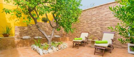 Un patio exterior con dos sillas de madera, un árbol pequeño y una maceta de piedra con plantas.
