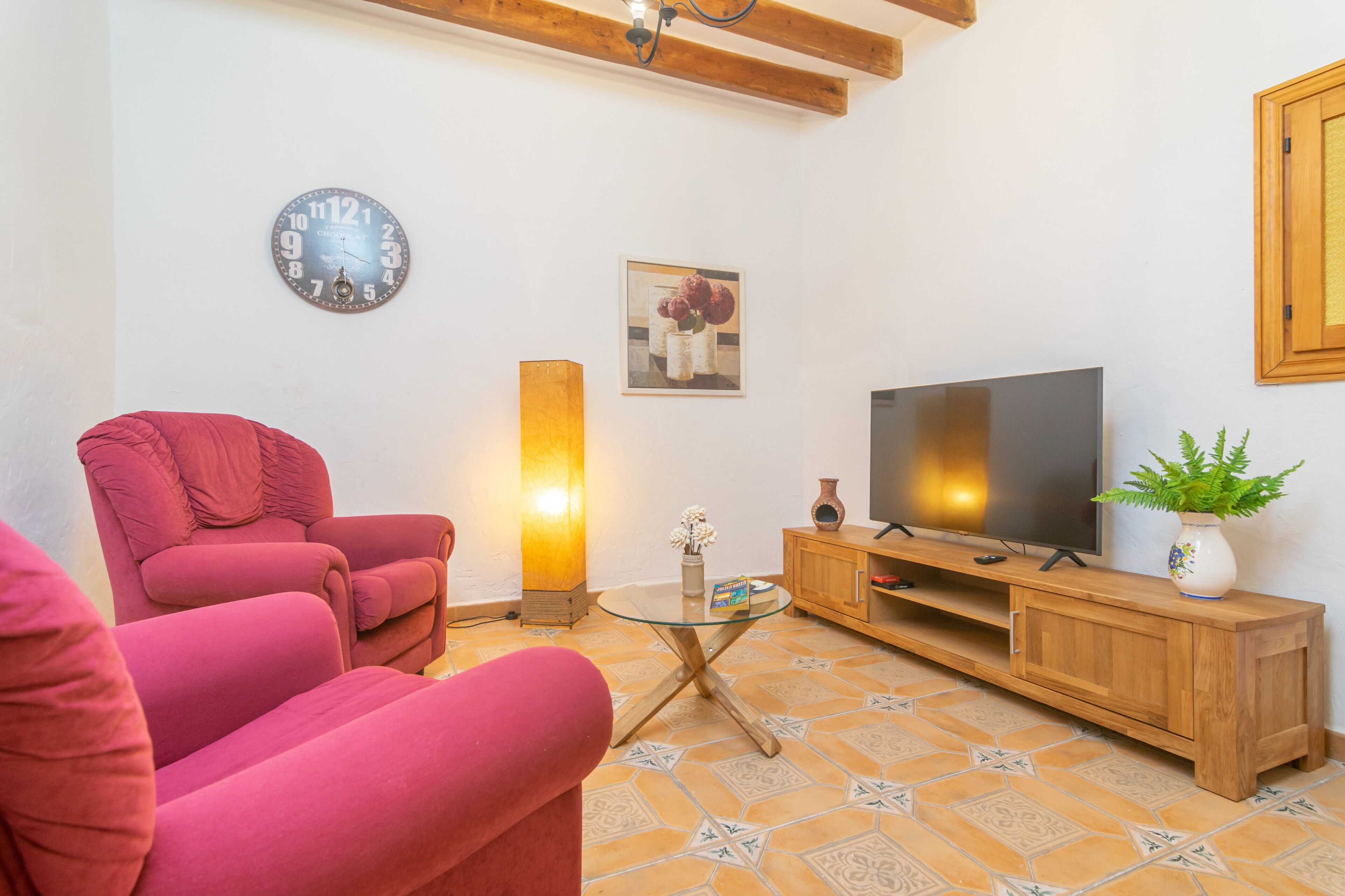 Una sala de estar con un sofá rojo, una mesa de centro de vidrio, un mueble de madera, un televisor de pantalla plana, un reloj de pared y un cuadro.