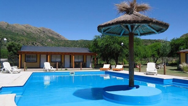 Une piscine avec des chaises longues, un parasol en chaume et un bâtiment doté de nombreuses fenêtres, le tout dans un cadre naturel.