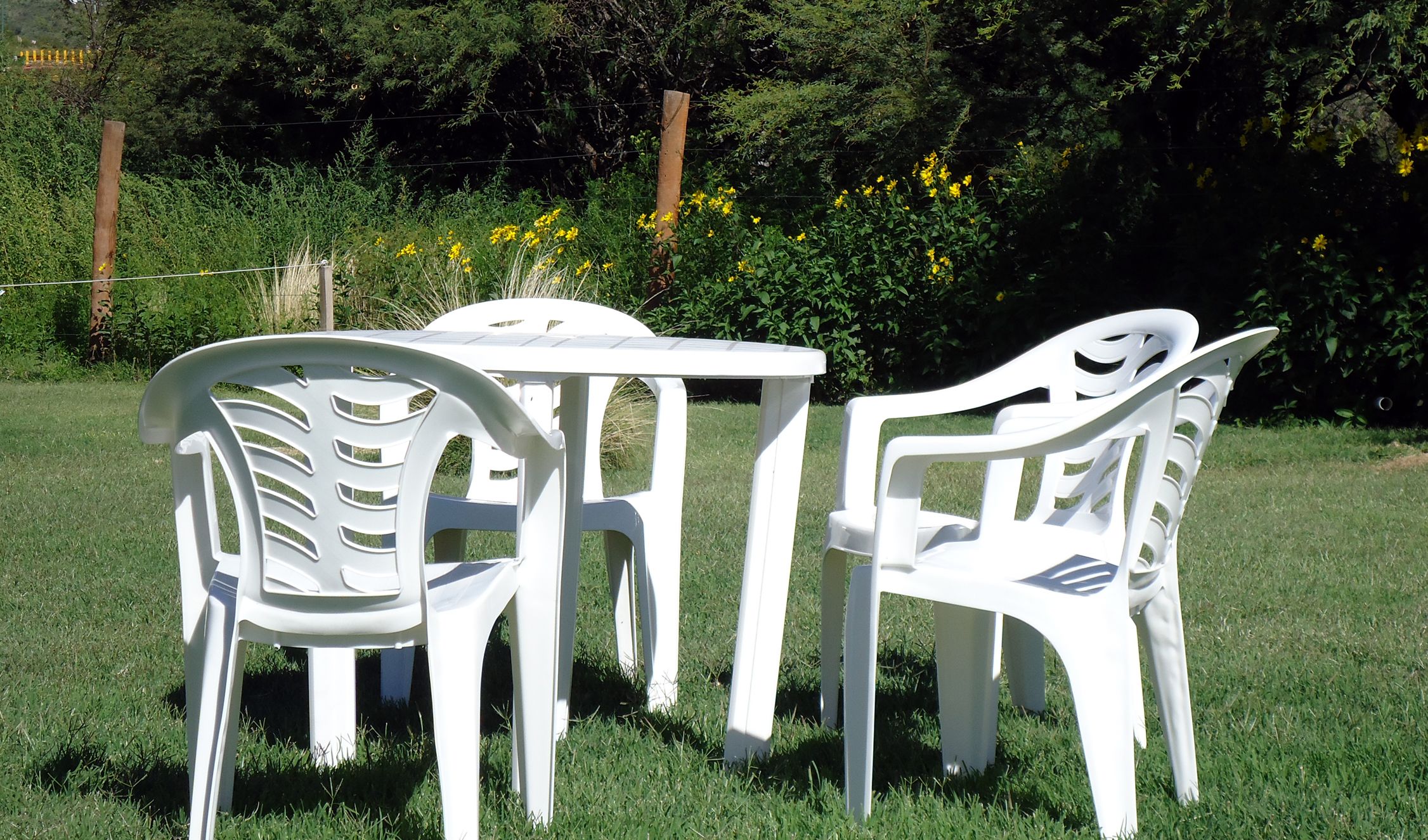 Una mesa blanca de plástico y cuatro sillas colocadas en un césped con una cerca y follaje al fondo.
