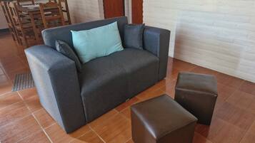A grey sofa with a light blue pillow, a brown ottoman, and a dining area with wooden chairs and a table.
