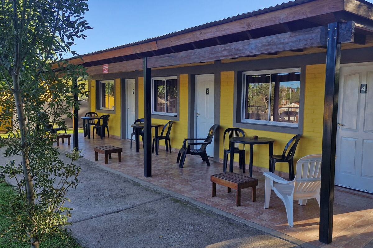 Un edificio con fachada amarilla, varias ventanas y un área de estar al aire libre cubierta con sillas y mesas de plástico.