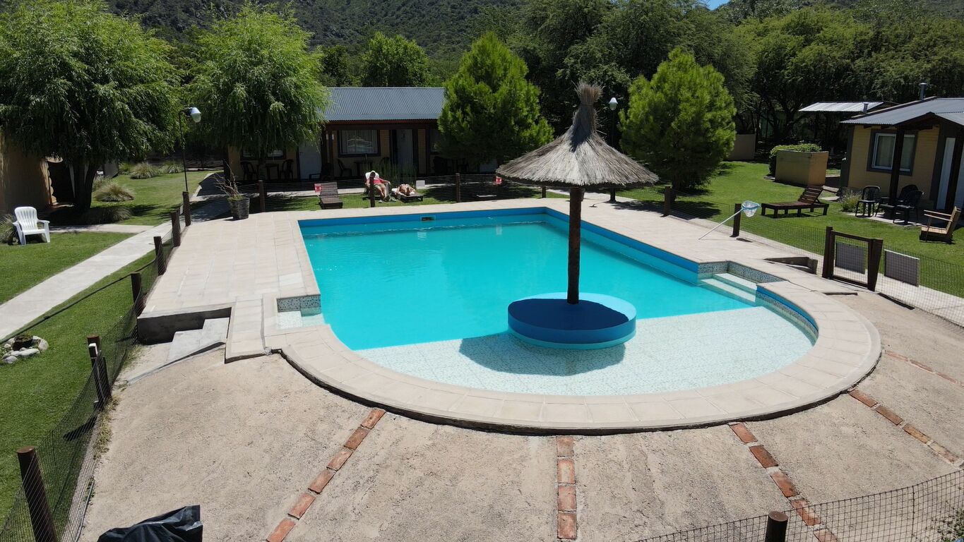 Una piscina con un deck circular, rodeada por una valla. Se observan sillones de descanso y un pequeño edificio al fondo.