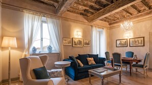 A cozy living room with a blue sofa, a white armchair, a round coffee table, and a dining table with chairs. There are framed pictures on the wall, a floor lamp, and a window with curtains. - Prestigious apartment with panoramic terrace, located in the center of Cortona. (Cortona)