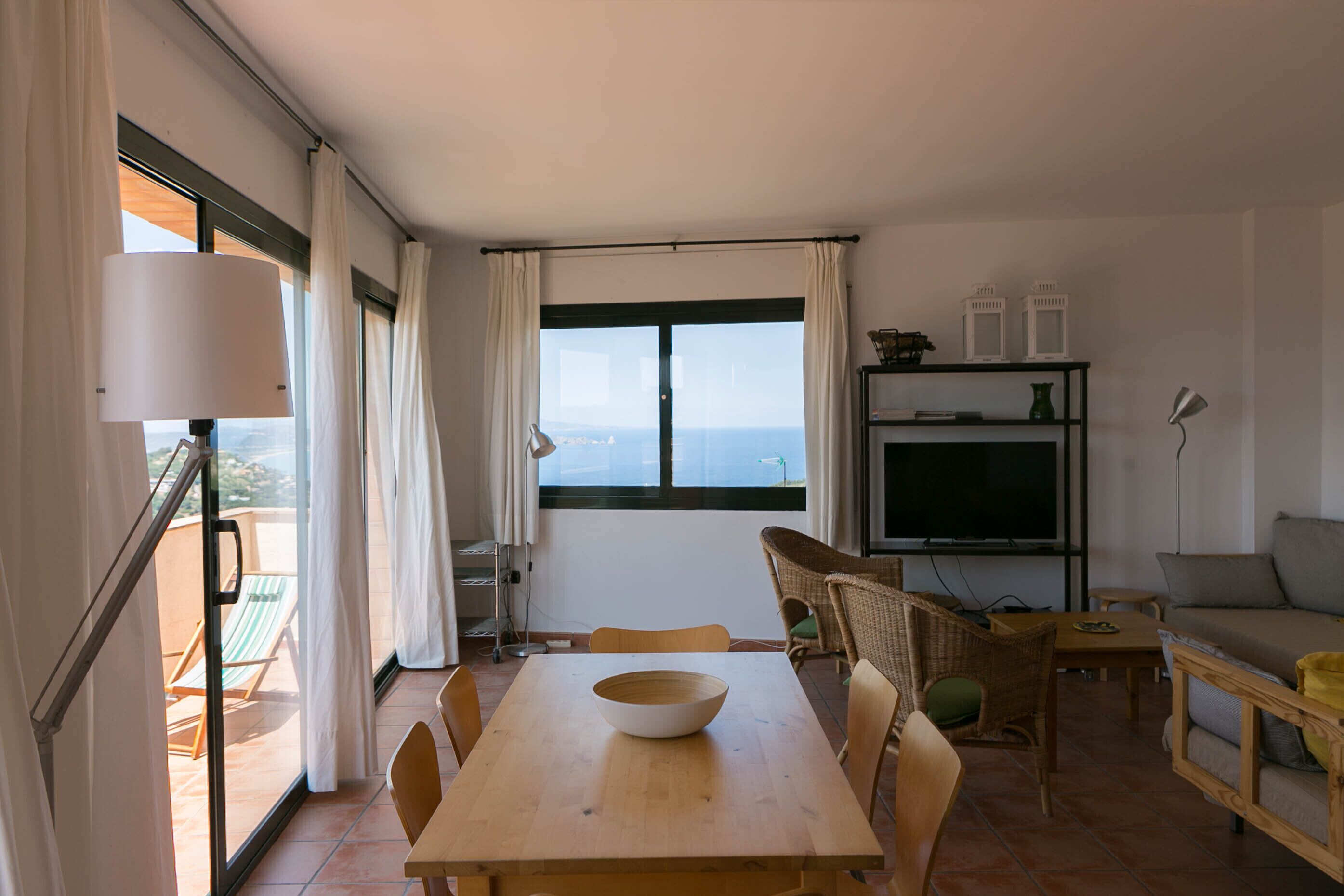 Un área de comedor con una mesa y sillas, un sofá, un televisor y vista al mar a través de un ventanal grande.