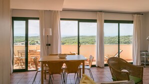 A dining area with a table and chairs, a wicker chair, and a view of a green landscape through large windows. - 2 bedroom apartment in Begur center. Sea views, terrace and pool (Ref:H09) (Begur)