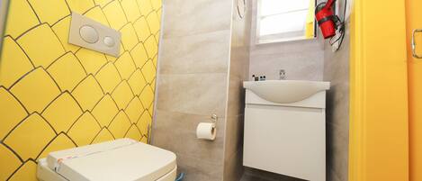 Un baño con pared de azulejos amarillos, inodoro blanco, lavamanos y zona de ducha.