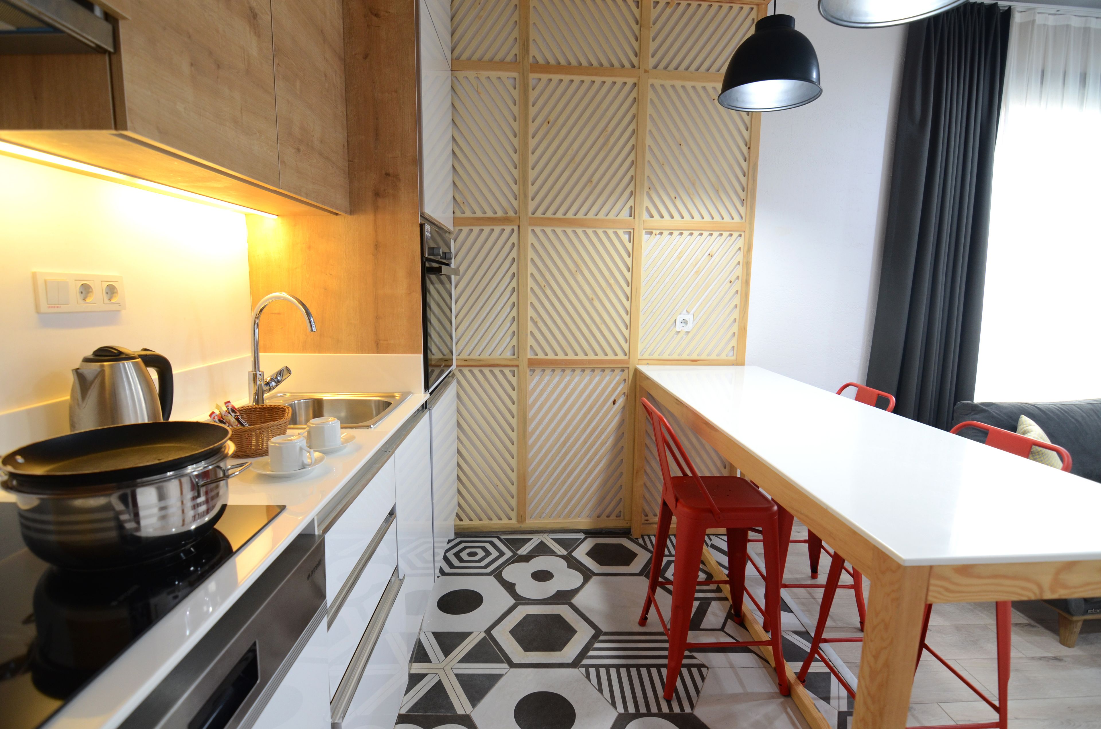 A modern kitchen with a white countertop, a stove, and a sink. There is a wooden partition with a geometric pattern, and a dining area with a white table and red chairs.
